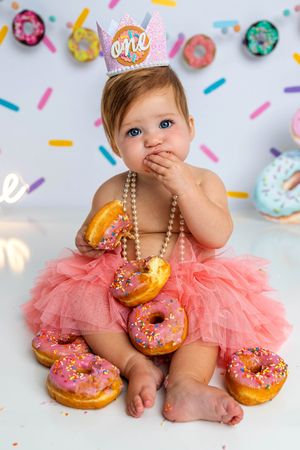 Baby girl in pink tutu and “one” crown sits surrounded by sprinkled donuts during first birthday cake smash photo session.