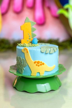 Blue dinosaur first birthday cake with yellow brontosaurus decoration and number one topper on green cake stand.