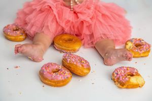 Baby in pink tutu dress sitting among pink frosted sprinkle donuts on white background.