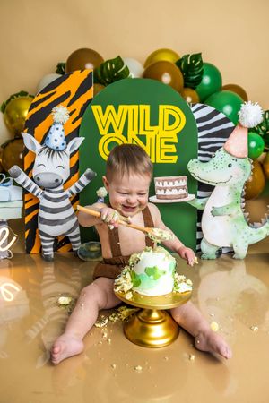 Baby smashing first birthday cake at Wild One safari themed party with zebra and dinosaur decorations and green gold balloons.