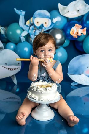 Baby enjoying first birthday cake smash with ocean theme decorations, sharks, and blue balloons in background.