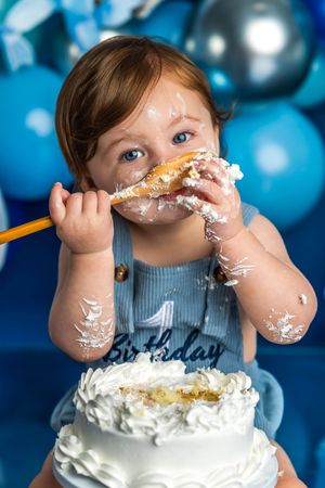 Messy baby celebrating first birthday smashing white cake, covered in frosting, blue balloons in background.
