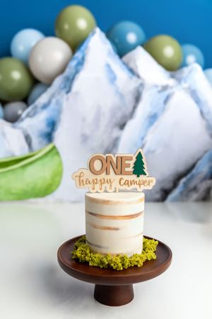 One Happy Camper first birthday smash cake on wooden stand with mountain backdrop and balloon decorations.