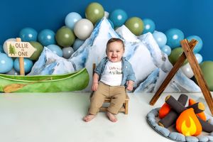 Baby boy smiling in camping-themed first birthday photo with mountains, balloons, canoe, and campfire decorations.