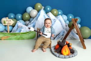 Baby boy smiles at camping-themed first birthday cake smash with mountains, balloons, canoe, and campfire decorations.