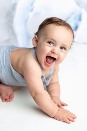 Happy baby in blue romper crawling on white floor, smiling with mouth open, bright studio setting.