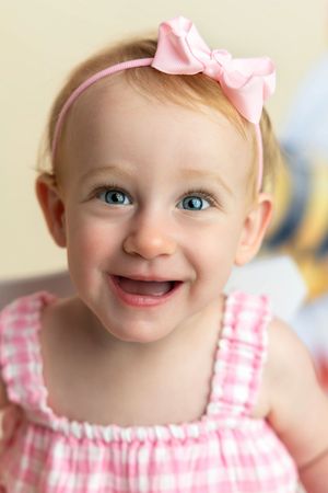 Smiling baby girl with blue eyes wearing a pink gingham dress and pink bow headband.
