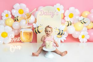 Baby girl sits with cake at a Daisy bee-themed first birthday party with floral balloon backdrop.