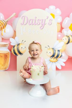 Baby girl smiling at her 1st birthday bee-themed cake smash with daisy decorations and pink balloon backdrop.