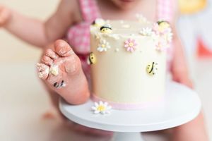 Baby's frosting-covered toes beside a decorated first birthday smash cake with bees and daisies on white stand.