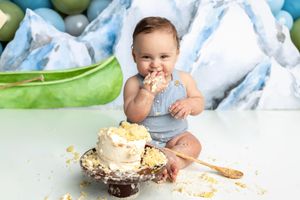 Baby in blue romper smashing first birthday cake, surrounded by crumbs, with balloon backdrop.