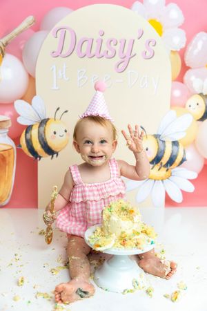 Happy baby girl in pink outfit smashing first birthday cake at Daisy's 1st Bee-Day party.