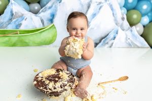 Baby enjoying first birthday cake smash session with mountain balloon backdrop, covered in white frosting.