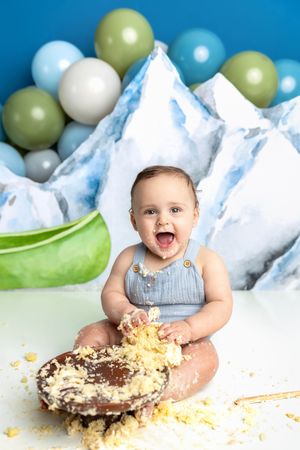 Baby smashing first birthday cake with mountains and balloons backdrop, covered in frosting, laughing joyfully.