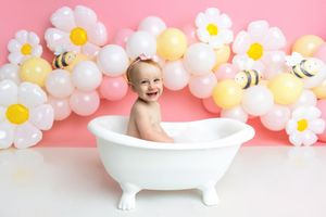 Baby smiling in white clawfoot tub with pink backdrop, daisy balloons, and bee decorations for a first birthday photoshoot.