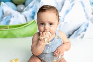 Cute baby boy in blue romper eating cake with his hands during a first birthday cake smash photoshoot.