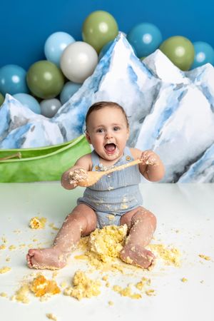 Excited baby covered in cake during first birthday smash cake photoshoot with mountain backdrop and balloons.