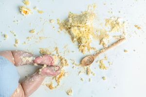 Baby hands covered in flour beside a wooden spoon on a light blue surface with scattered flour.