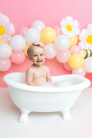 Smiling baby in a white clawfoot tub with pink balloon daisy backdrop for a first birthday photo shoot.