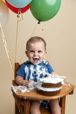 Happy baby boy celebrates first birthday smashing white frosted cake in wooden high chair with colorful balloons.