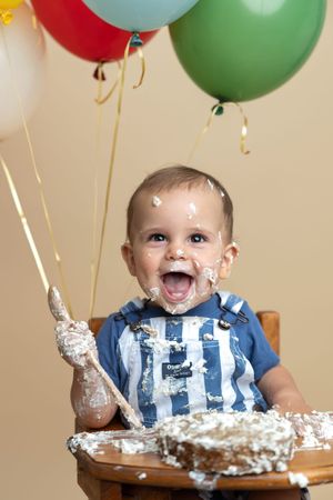 Happy baby covered in cake frosting celebrates first birthday in high chair with colorful balloons.