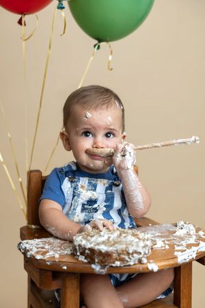 Baby in high chair celebrates first birthday smashing cake, covered in frosting, with colorful balloons in background.