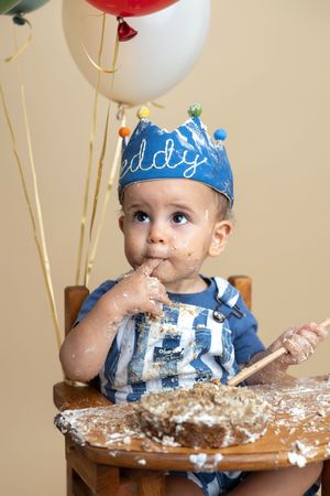 Baby boy in blue crown smashes first birthday cake in high chair, surrounded by colorful balloons.