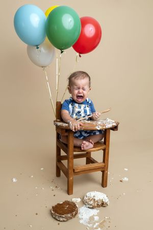 Crying baby in high chair with smashed birthday cake on floor, colorful balloons tied to chair, cake smash photo session.