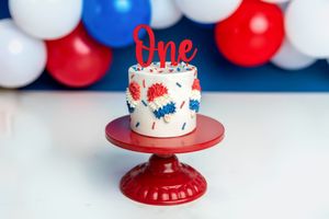 White first birthday smash cake with red and blue comic-style decorations and 'One' topper on red cake stand.