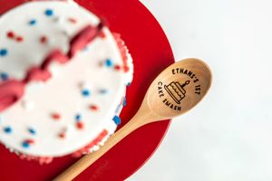 Personalized wooden spoon engraved with cake design next to a red, white, and blue sprinkle cake on a red plate.