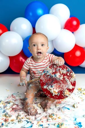 Baby boy smashing patriotic red cake at first birthday party with colorful balloons backdrop.