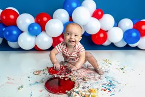 Baby smiling during first birthday cake smash with red, white, and blue balloons and patriotic decorations.