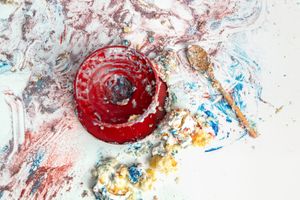 Red ceramic bowl with spoon on a colorful paint-splattered white surface with blue, red, and yellow hues.
