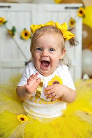 A joyful baby girl in a yellow tutu and sunflower onesie claps her hands, wearing a big yellow bow on her first birthday.
