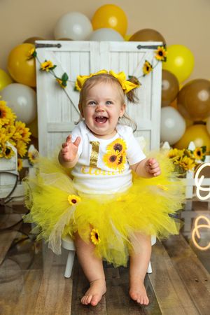 Happy baby girl in yellow tutu and sunflower outfit celebrates first birthday surrounded by gold and silver balloons.