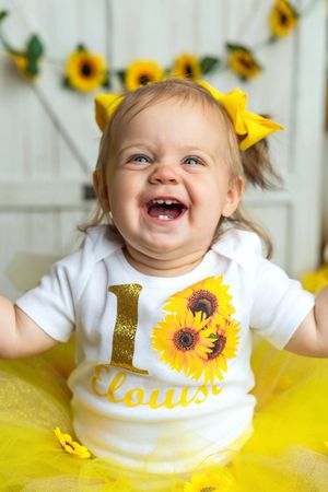 Baby girl Elouise laughing in sunflower birthday outfit with yellow tutu and bow for 1st birthday photo.