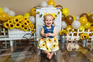 Smiling toddler girl in sunflower dress sits on white chair surrounded by yellow balloons and sunflowers at first birthday.