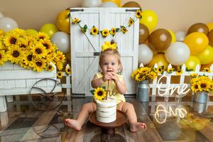 Baby girl in yellow outfit celebrates first birthday with sunflower cake smash, surrounded by yellow balloons and sunflower decorations.