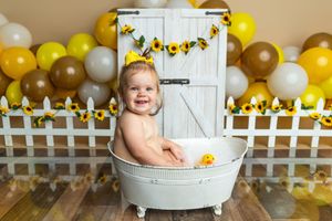 Smiling baby in a white bathtub surrounded by yellow, white, and brown balloons with sunflower decorations.