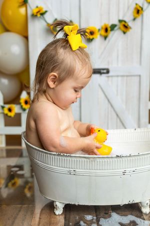 Baby girl with yellow bow sits in vintage tub holding rubber duck, surrounded by sunflower decorations and balloons.