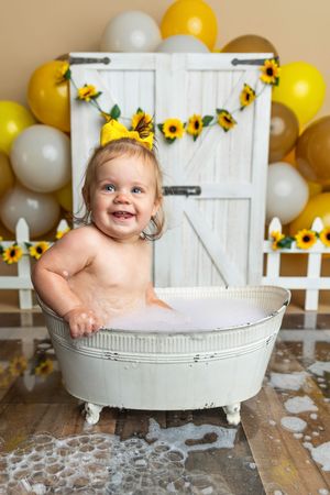 Smiling baby girl in a bubble bath tub with sunflower decorations and yellow balloons for a first birthday photo shoot.