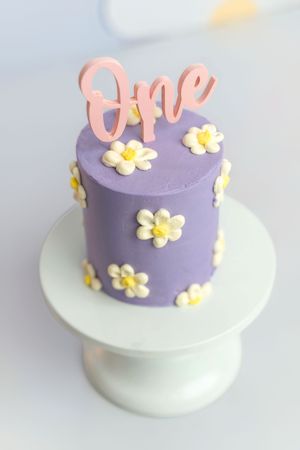 Purple first birthday smash cake with white daisy decorations and pink "One" topper on white cake stand.