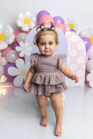 Cute baby girl in purple ruffle romper stands barefoot before floral balloon backdrop at first birthday party.