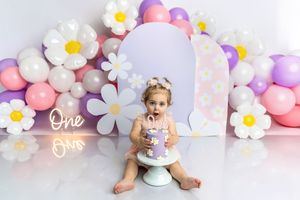 Baby girl smash cake photoshoot with daisy balloons, purple cake, and neon One sign for first birthday.