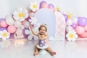 Baby girl smiling at her first birthday cake smash with daisy balloons and pastel decorations.