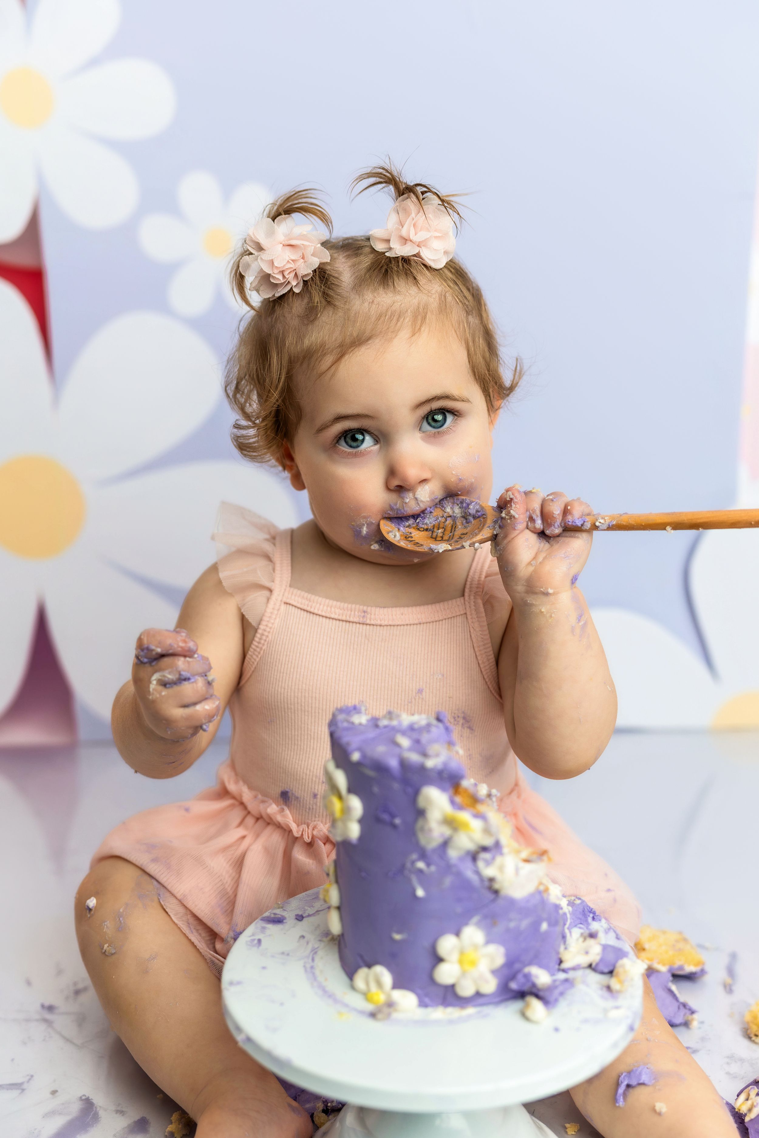 Toddler girl in pink tutu eating cake with spoon at first birthday cake smash with purple floral decorated cake.