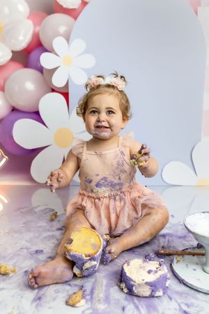 Smiling baby girl in pink tutu celebrates first birthday smash cake session with daisy and balloon backdrop.