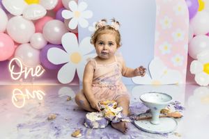 Baby girl in pink dress smashes first birthday cake surrounded by daisy balloons and One neon sign.