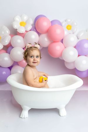 Baby girl sits in white clawfoot tub holding yellow duck, surrounded by pink and purple balloon arch with daisy decorations.