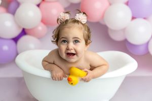 Smiling baby girl in white bathtub with rubber duck, pink and purple balloon backdrop, professional photo.